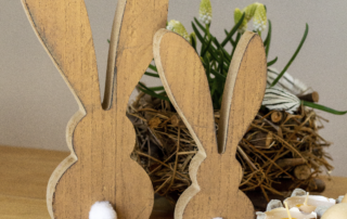 Zwei Deko-Osterhasen aus Holz stehen vor einem Osterstrauch auf dem Tisch.