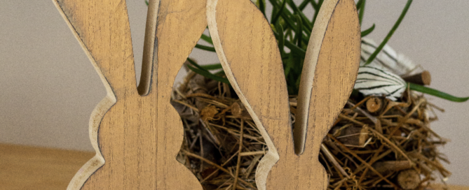 Zwei Deko-Osterhasen aus Holz stehen vor einem Osterstrauch auf dem Tisch.