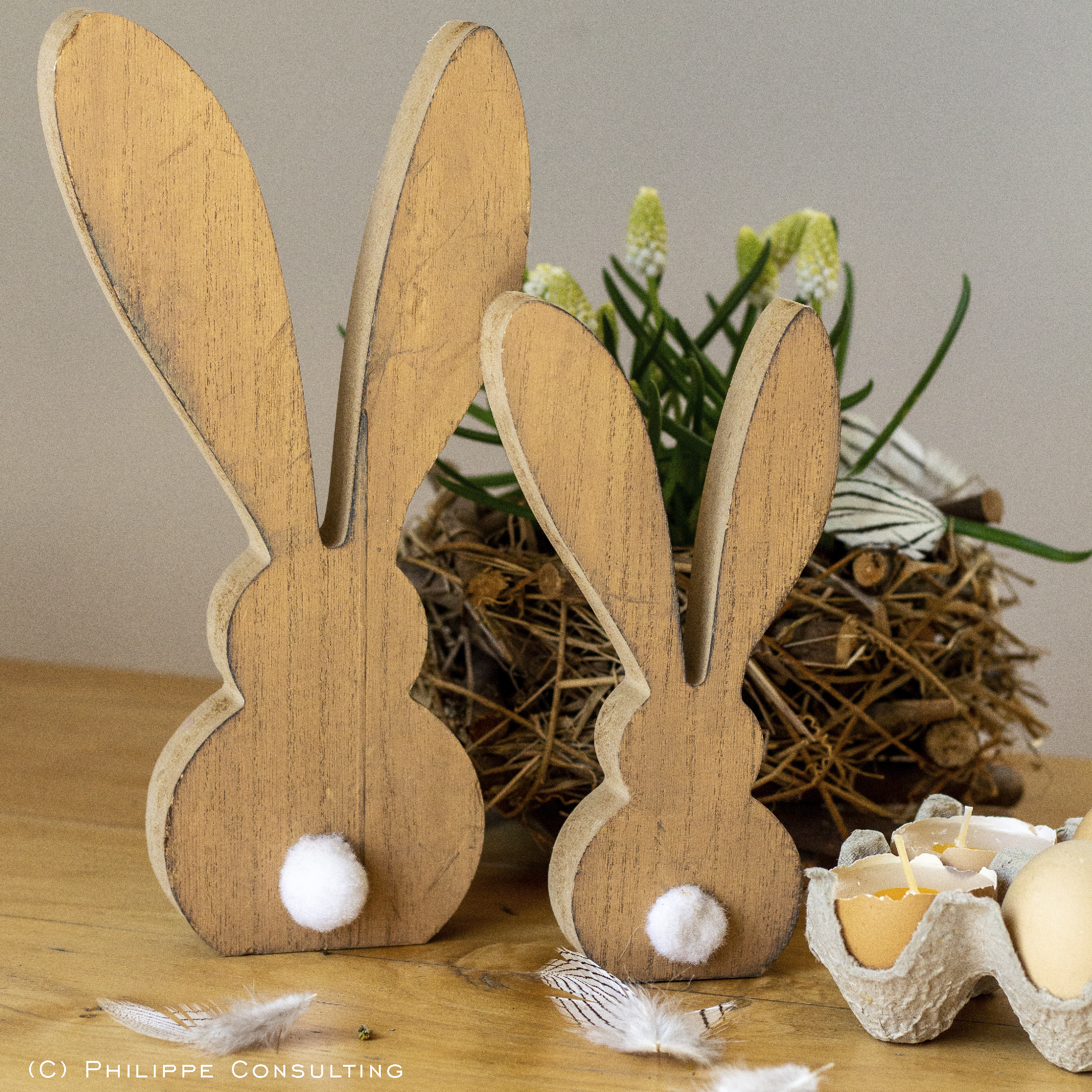 Zwei Deko-Osterhasen aus Holz stehen vor einem Osterstrauch auf dem Tisch.
